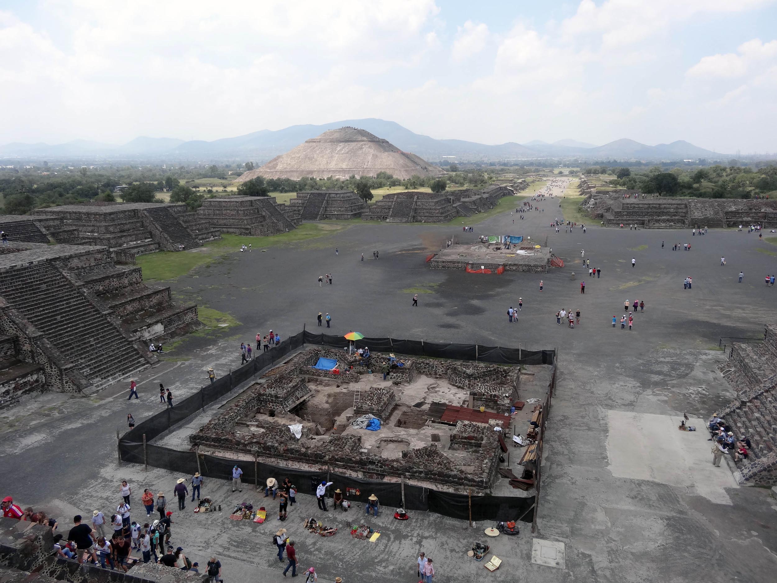 In the center of the city of Teotihuacan, people crowd around an archaeological site. Inside the site we can see remnants of brick structures. The site has a black fence around it.