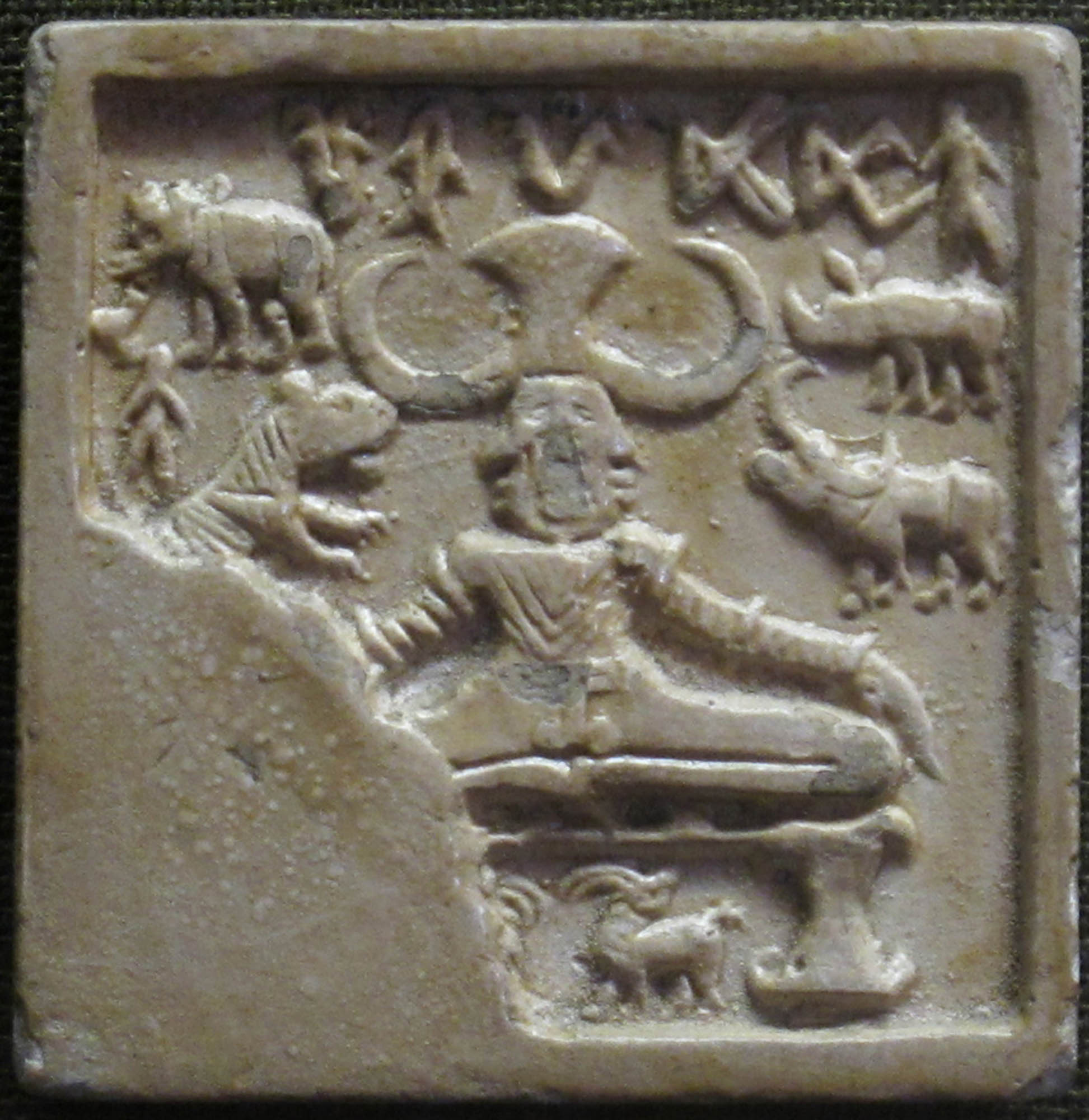 Small, square, stone carving with a male figure in a yogic position and surrounded by animals and written text.