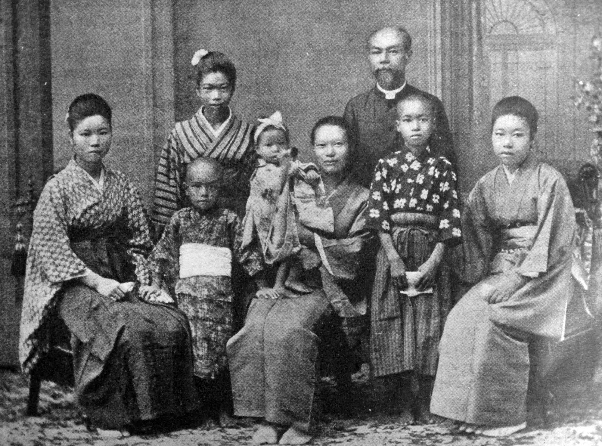A large family of nine poses for a photograph. Most wear traditional Japanese robes but a man wears the collar and coat of a Christian pastor.