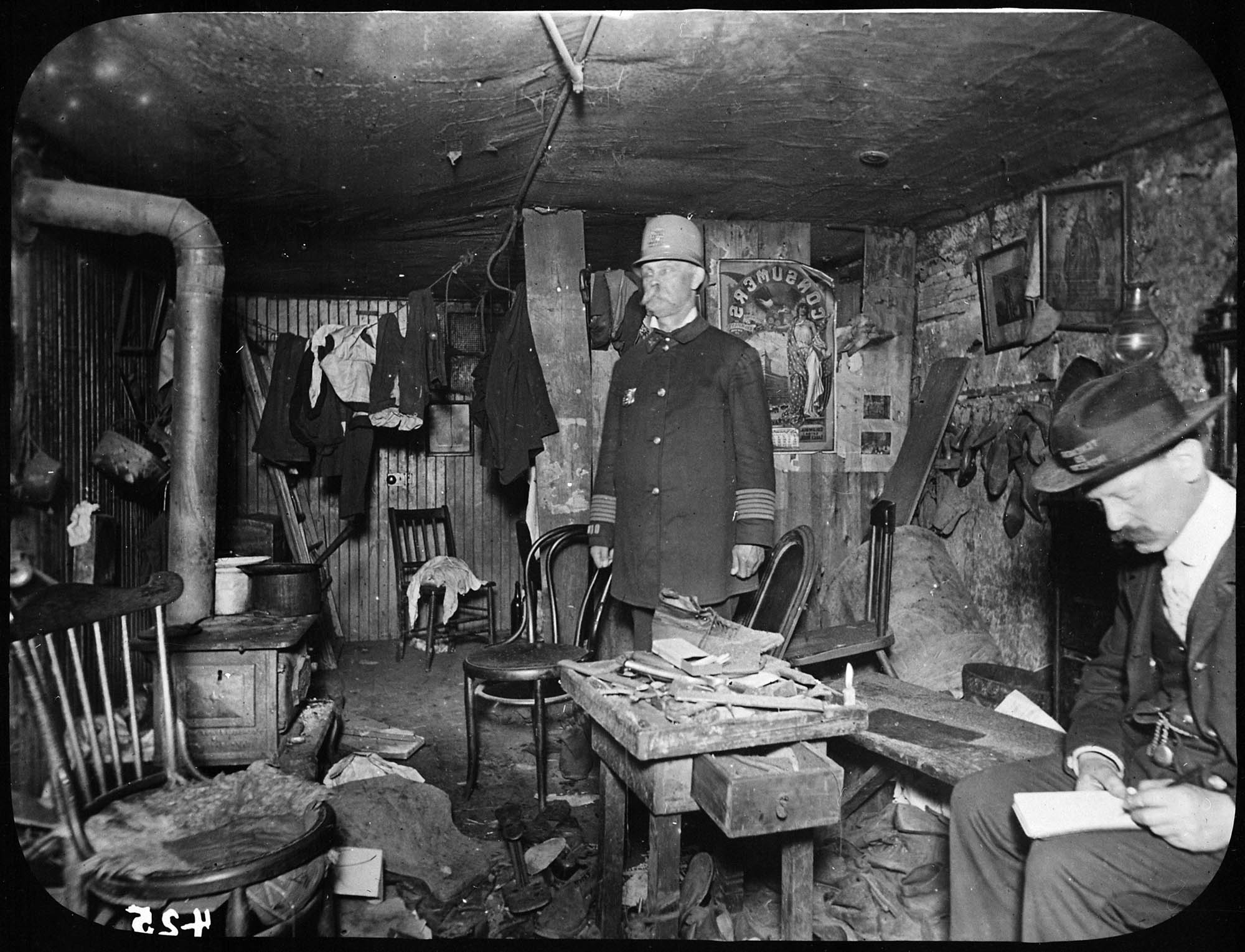 A policeman and a city official inspect a run-down, dirty, and cluttered basement apartment.