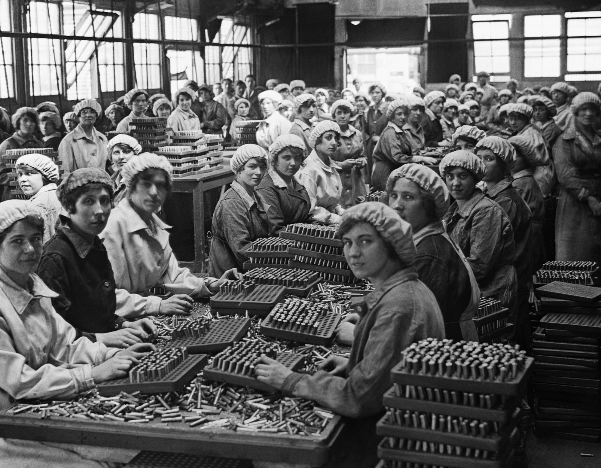 A black-and-white photograph of a large group of women in factory uniforms and caps assembling ammunition at long tables inside a warehouse.