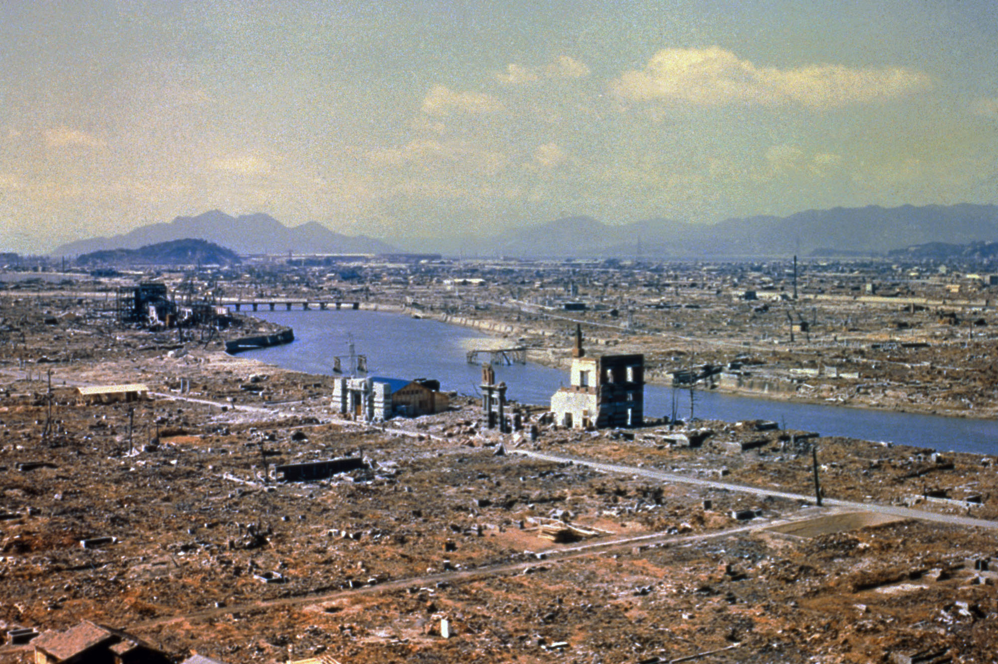 Aerial photograph of Hiroshima showing widespread devastation after the atomic bombing. Rubble stretches across the city with only a few buildings left standing near a river, and mountains rise in the distance.