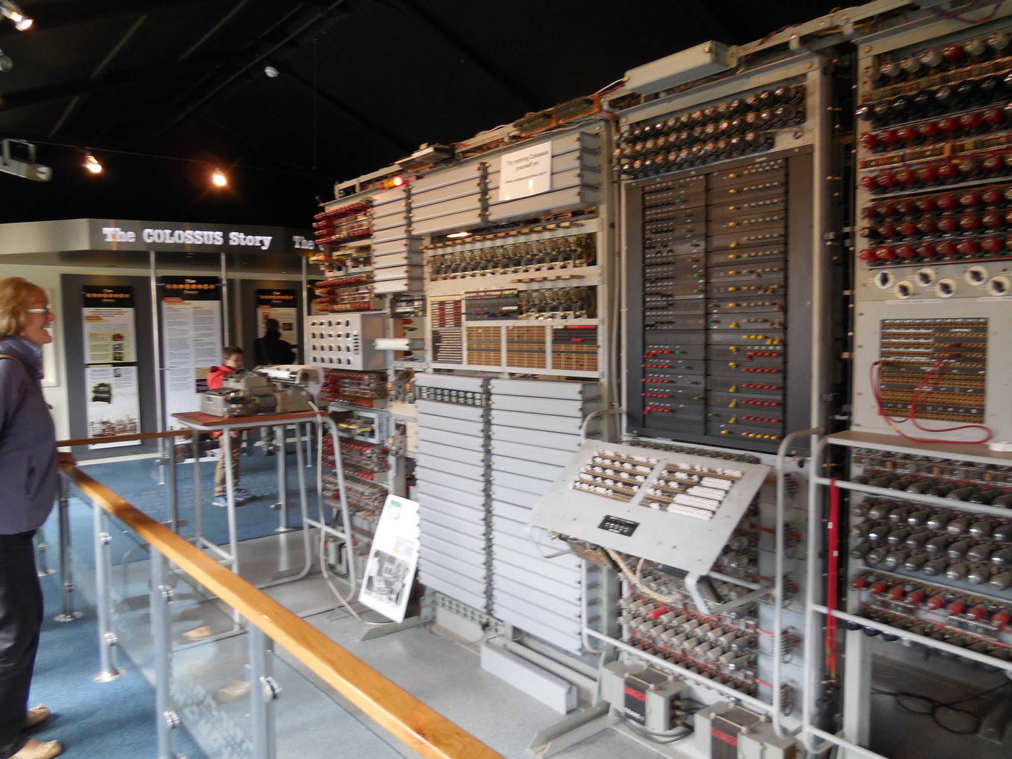 A museum display of Colossus, an early digital computer, with large panels filled with switches, wires, and dials extending along a wall. A visitor stands nearby behind a railing.