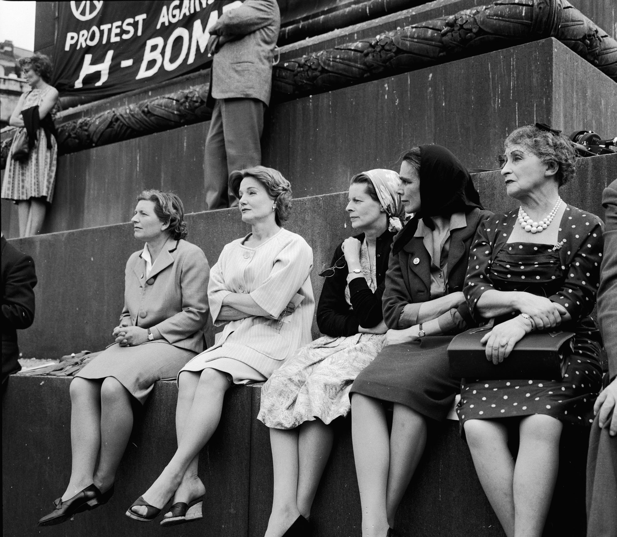 A black-and-white photograph of a group of women sitting on a stone ledge, dressed in coats and dresses, with a large protest banner behind them reading “PROTEST AGAINST H-BOMB.”