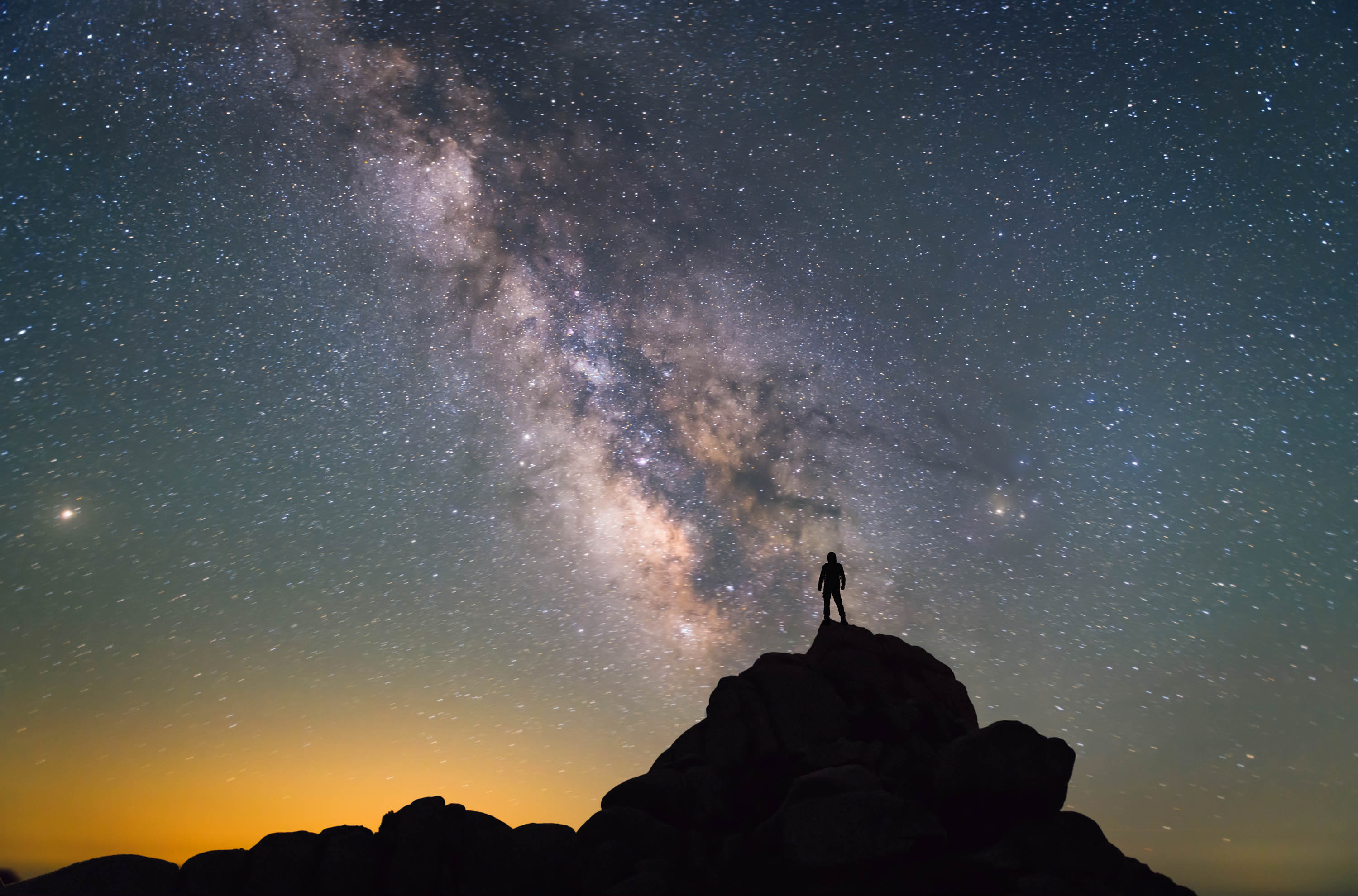 Silhouette of a person standing on a rocky hill beneath the glowing Milky Way in a star-filled night sky.