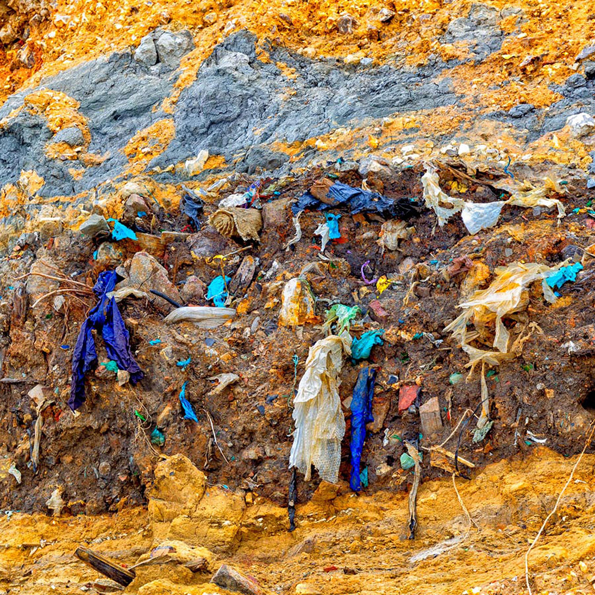 Layer of exposed earth showing buried plastic waste and textiles embedded in soil and rock.
