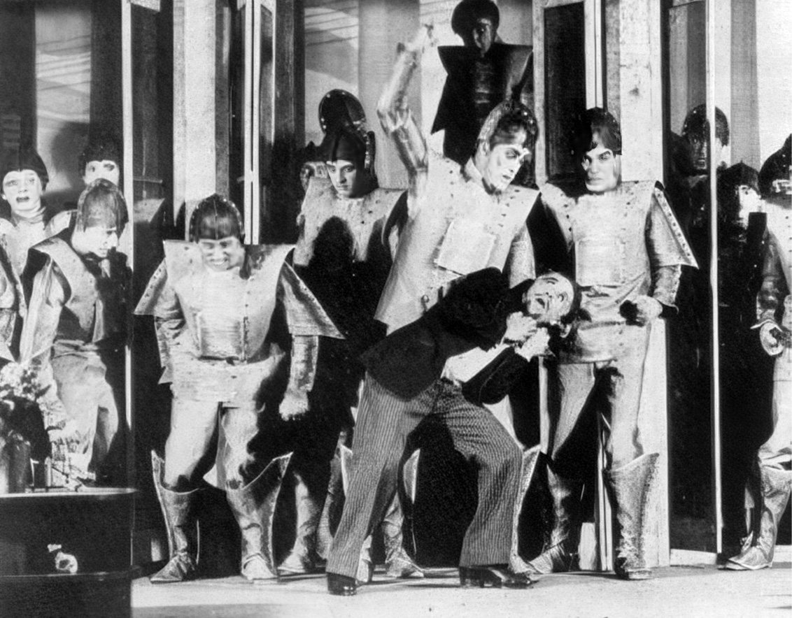 A screenshot from a 1920s film of people dressed as robots attacking a man in a suit.