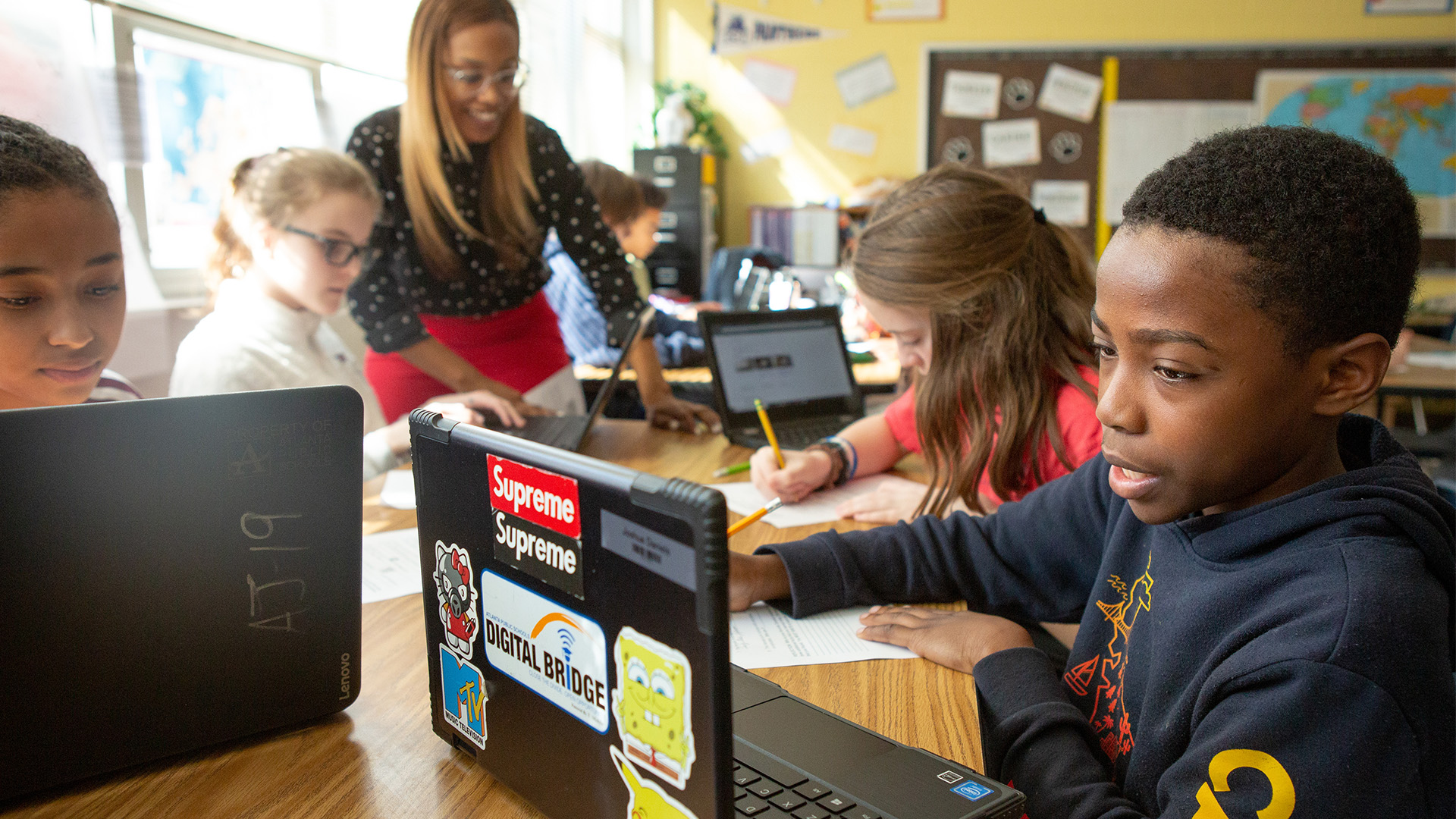 Five young students work with laptops while a teacher watches on.