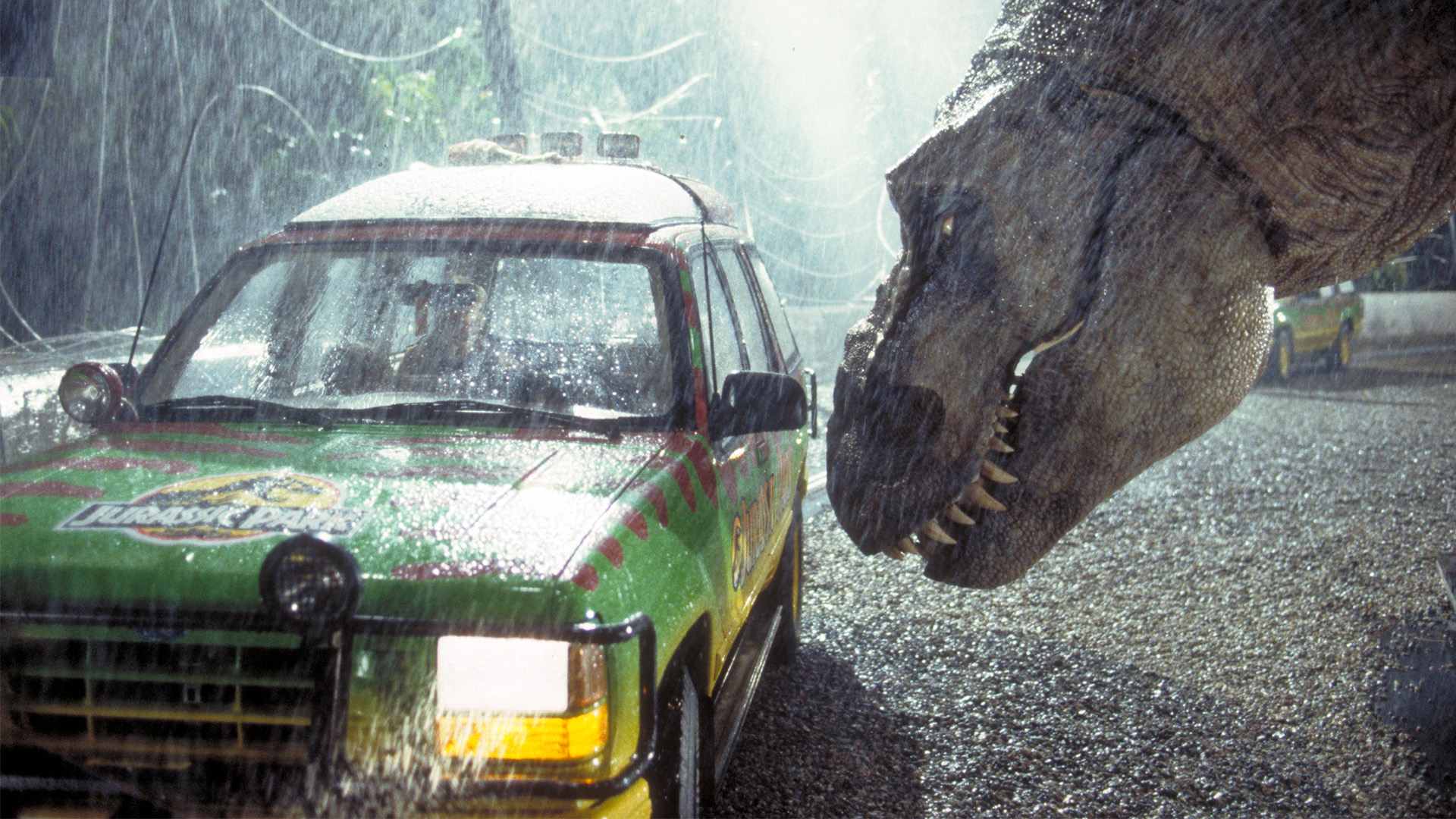 A screenshot from the original Jurassic Park film featuring a Tyrannosaurus in the rain peering into the window of a car.