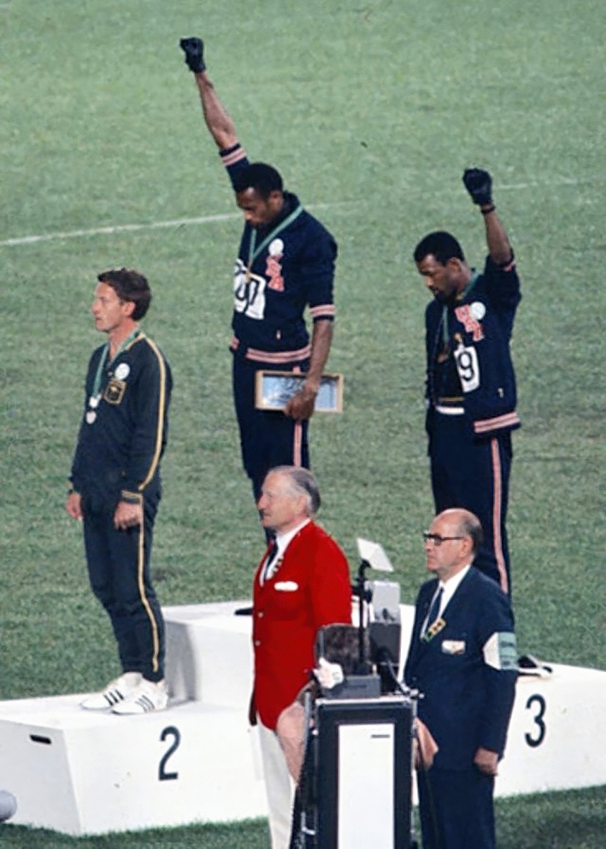 Two American athletes stand on a podium at the 1968 Olympics. Each wears one black glove and have hands raised in protest.