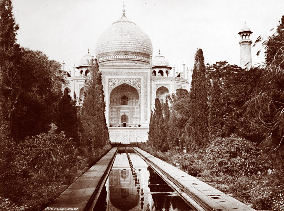 A historic sepia-toned photograph of the Taj Mahal, with its white marble dome and arched façade reflected in a long rectangular pool, framed by trees and garden pathways leading toward the monument.