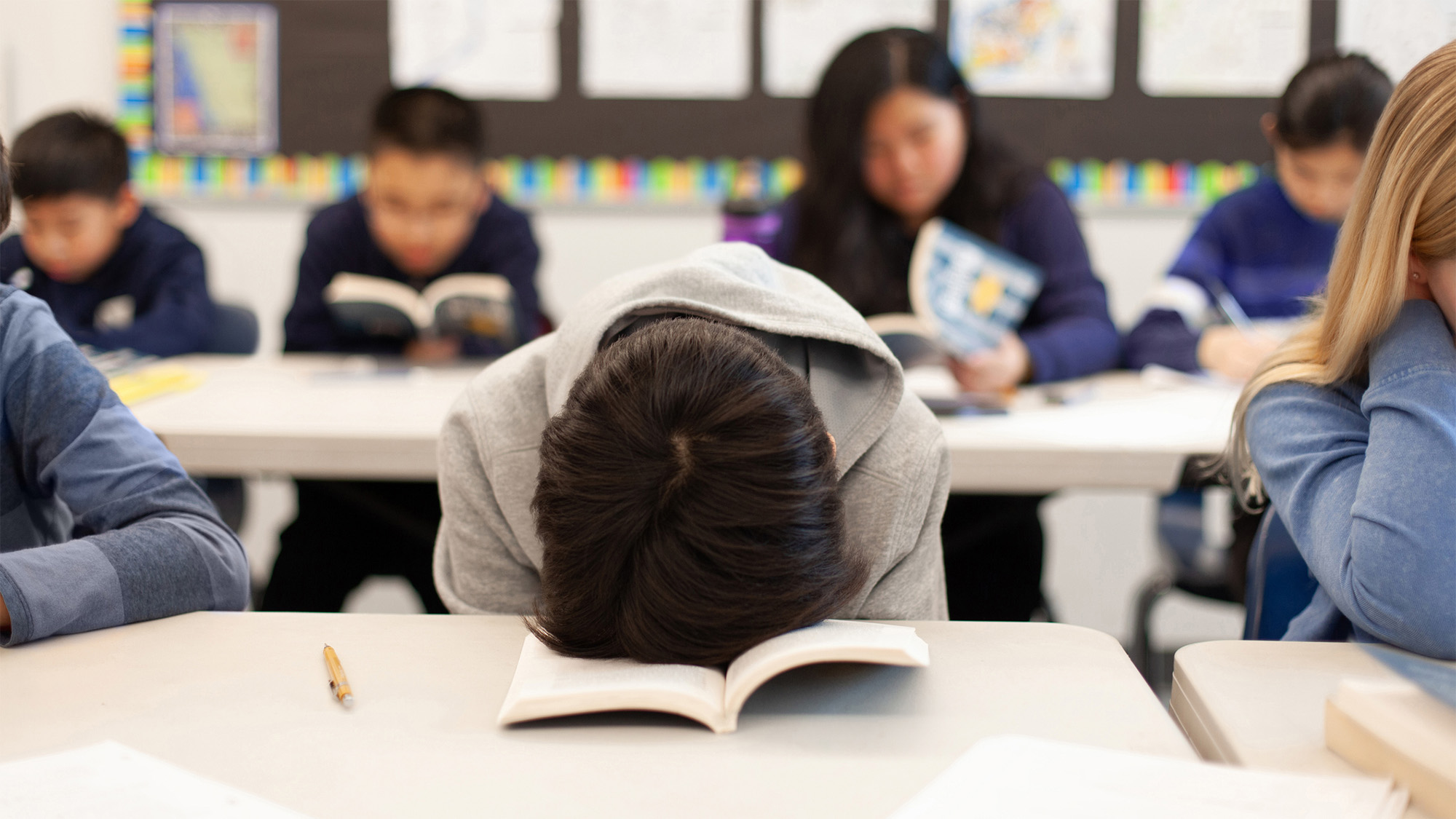 A student rests their head on an open book at a classroom desk while other students sit nearby reading quietly in the background.