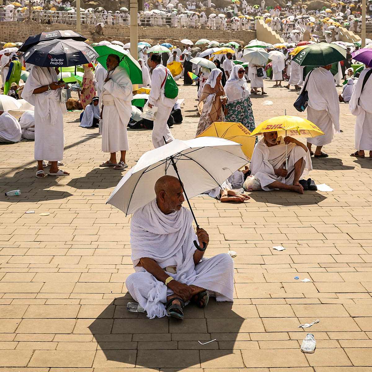 A man sits on the ground holding an umbrella to shelter from the hot sun. A crowd of people stand behind some also umbrellas.