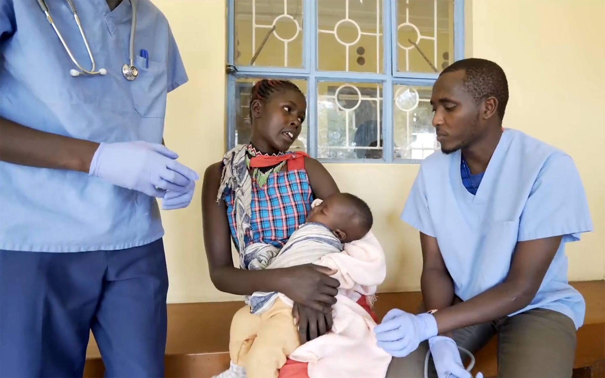A new mother cradles a newborn in her arms. The baby is wrapped tight in blankets and sleeping. The mother is speaking with healthcare workers at a health clinic.