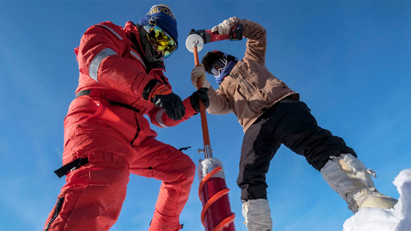 Low angle view of two scientists wearing warm clothes and using tools to core ice in Utqiavik, Alaska.