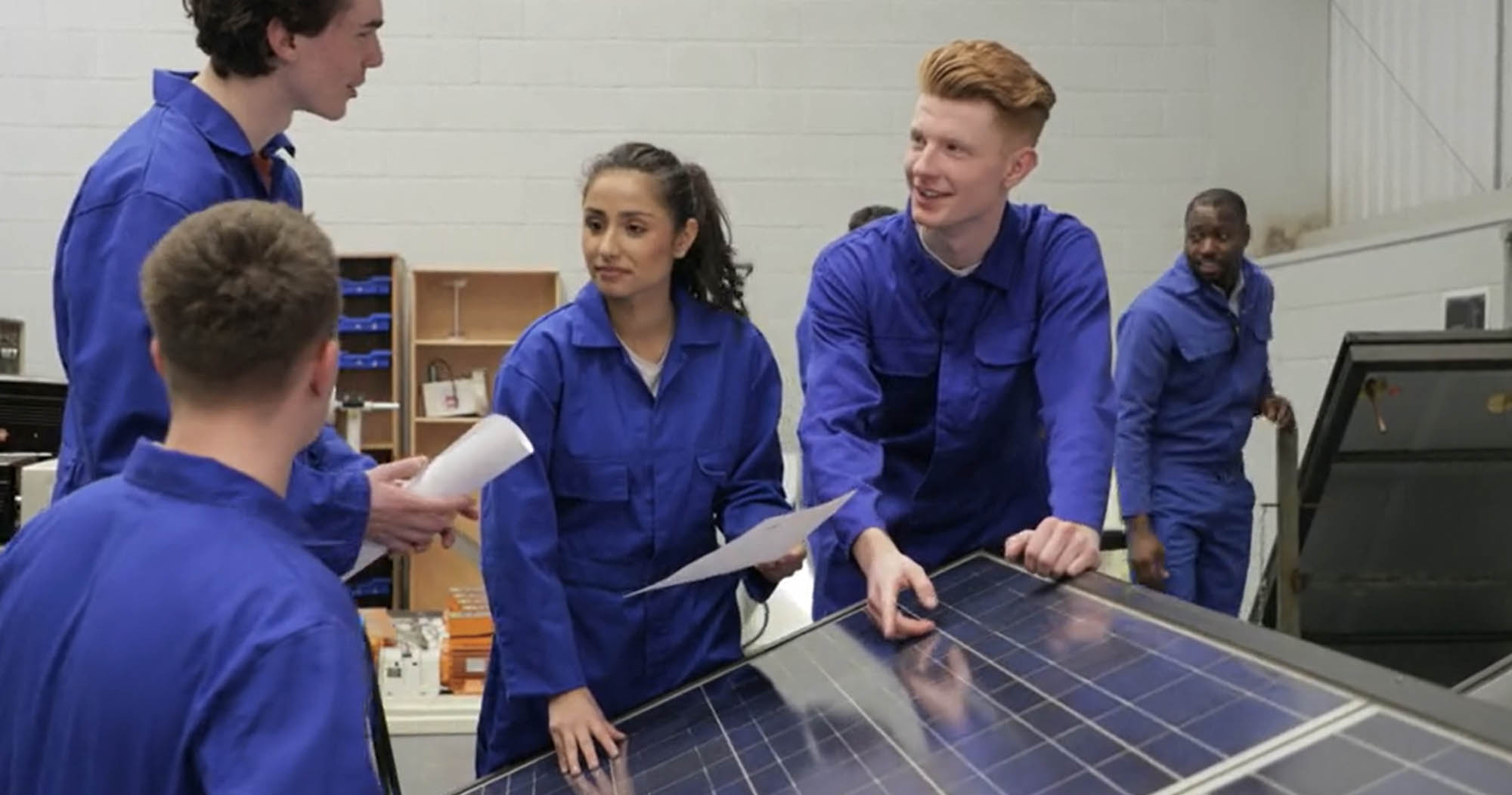 Multiple young students wearing blue lab coats in discussion about solar energy.