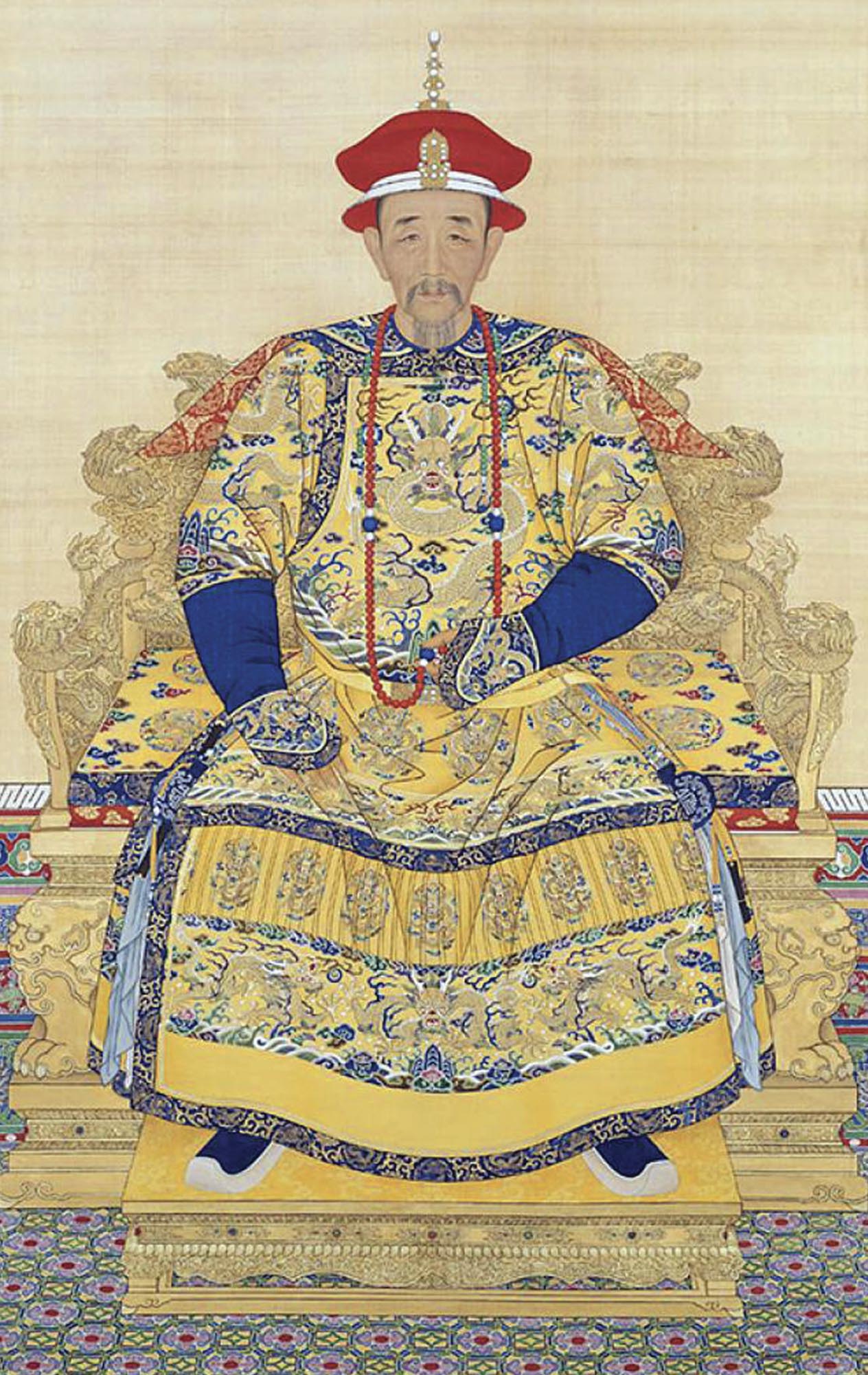 Painting depicts the Kangxi Emperor sitting in a very ornate chair or throne, wearing detailed and ornate clothing in shades of yellow and blue.