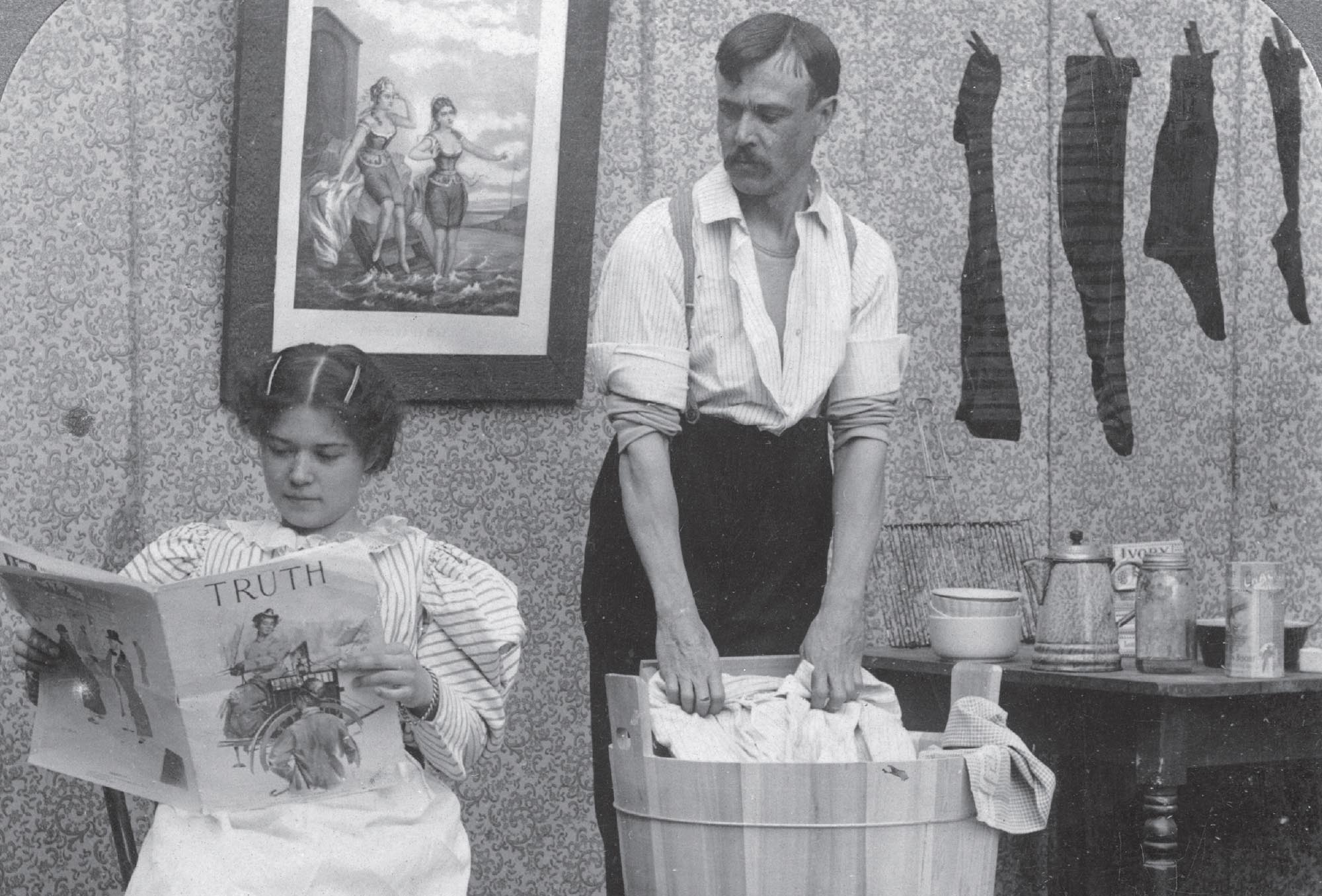 Black and white photo of a man, with his sleeves rolled up, doing the washing while his wife relaxes reading a newspaper.