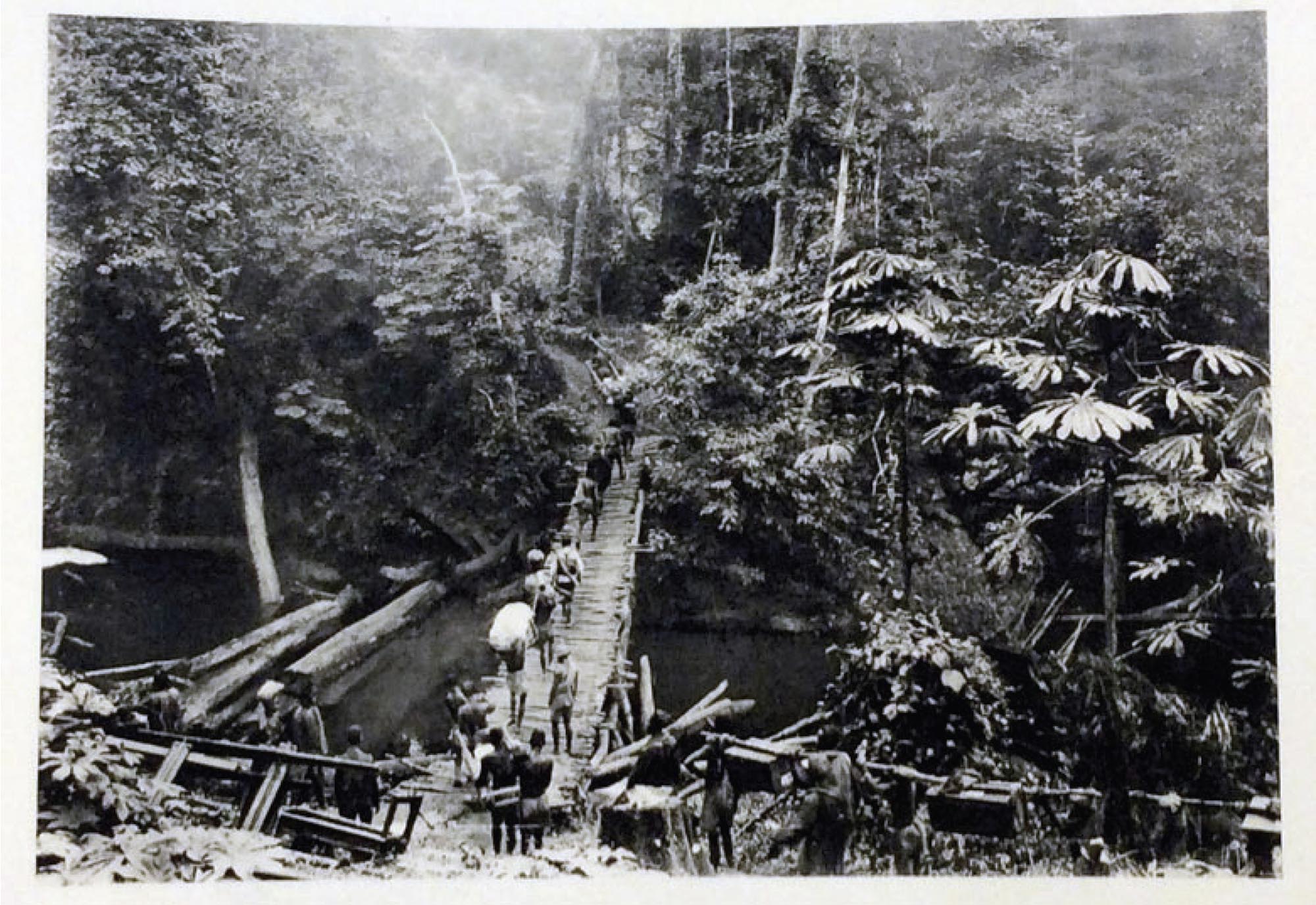 Forced laborers carry supplies across a suspended wooden bridge over a river.