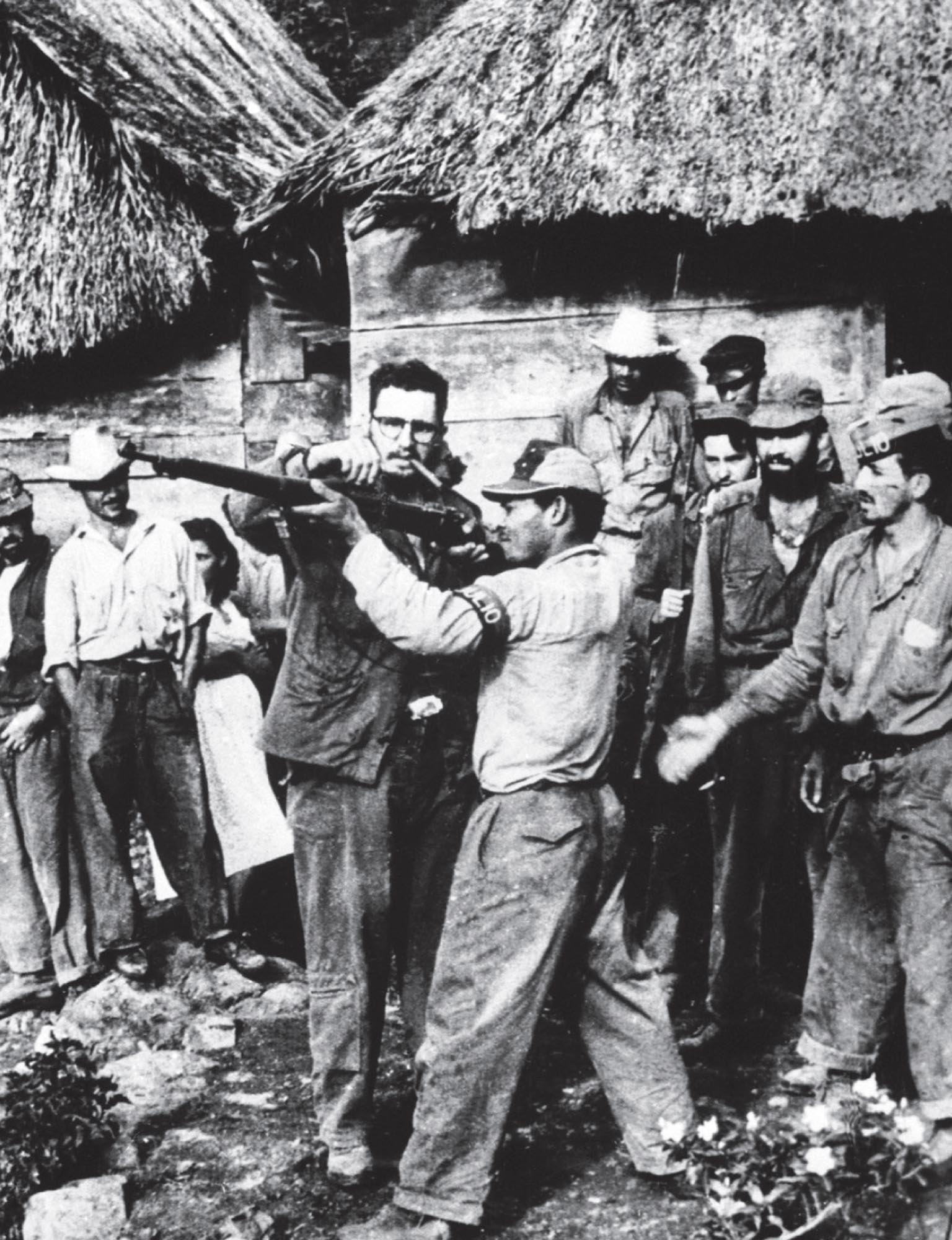 Fidel Castro giving arms training to a group of guerilla fighters. Group stands in front of simple thatched-roof cabins.