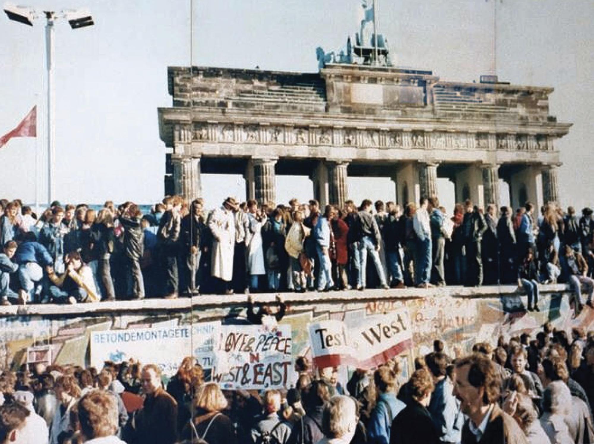 Photo of a crowd gathered on and around the Berlin wall. Some carry signs in support of peace between East and West Germany.