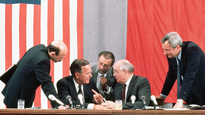 Photo of President George H.W. Bush, Soviet leader Mikhail Gorbachev, and their associates sitting behind a table, deep in conversation. Behind them on the wall are American and Soviet flags.