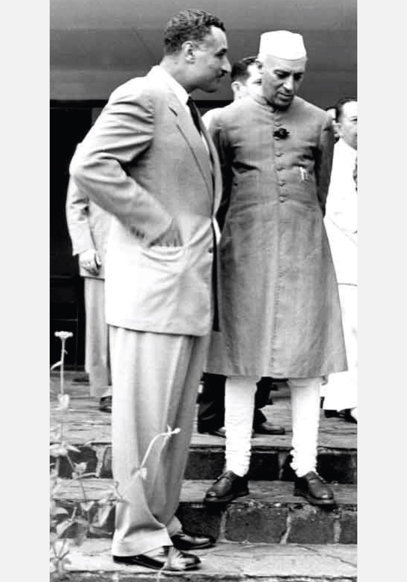 Two men standing together in conversation, the man in a suit has hands in his pockets.