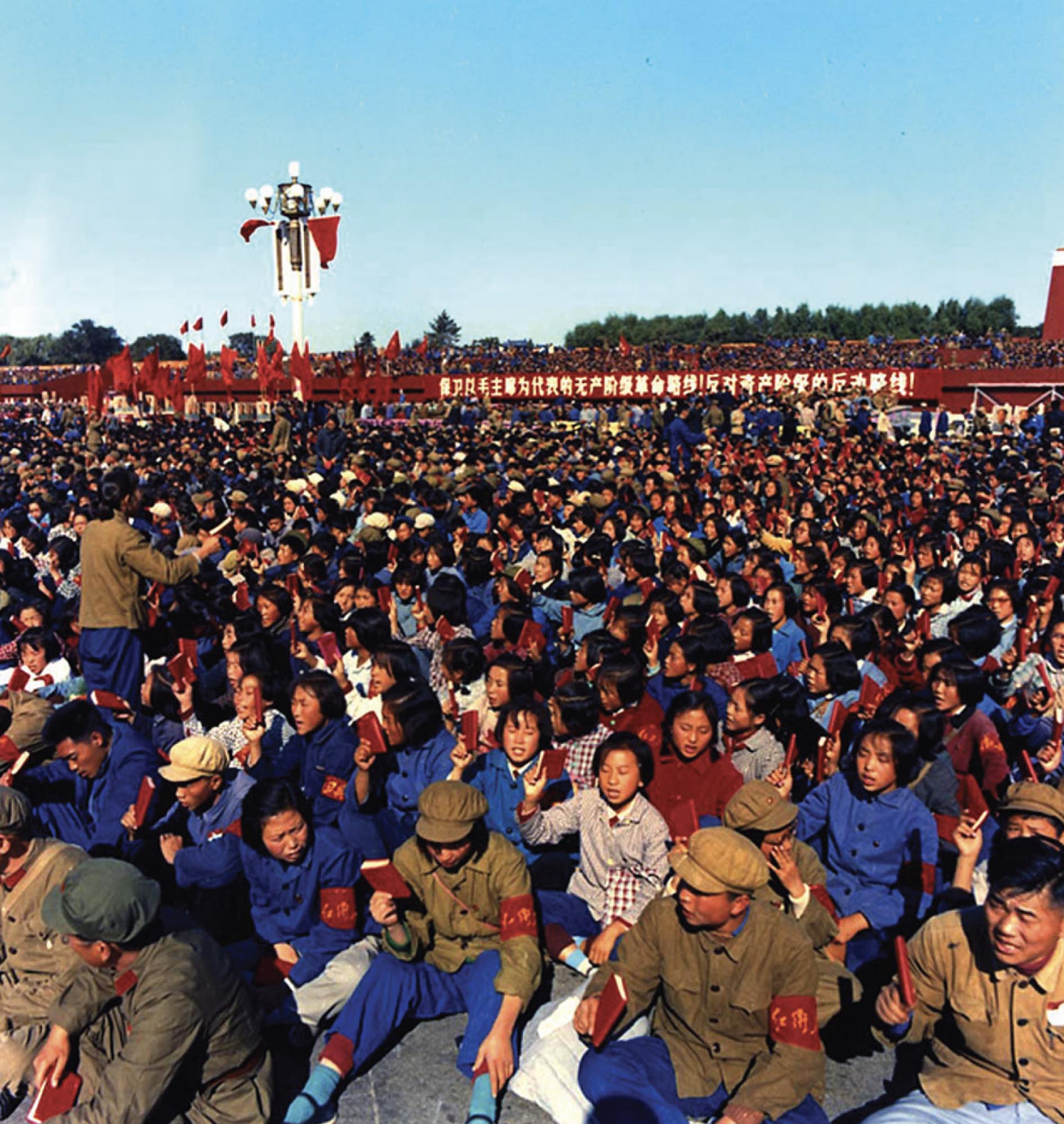 Photo of a large crowd of young students sitting on the ground holding small, red books in their hands. Each student is wearing a red armband.  