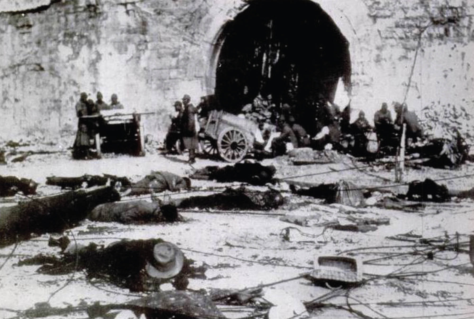 Photo of massacred civilians lying dead on the street. In the background, bodies are being piled onto a large wooden cart.