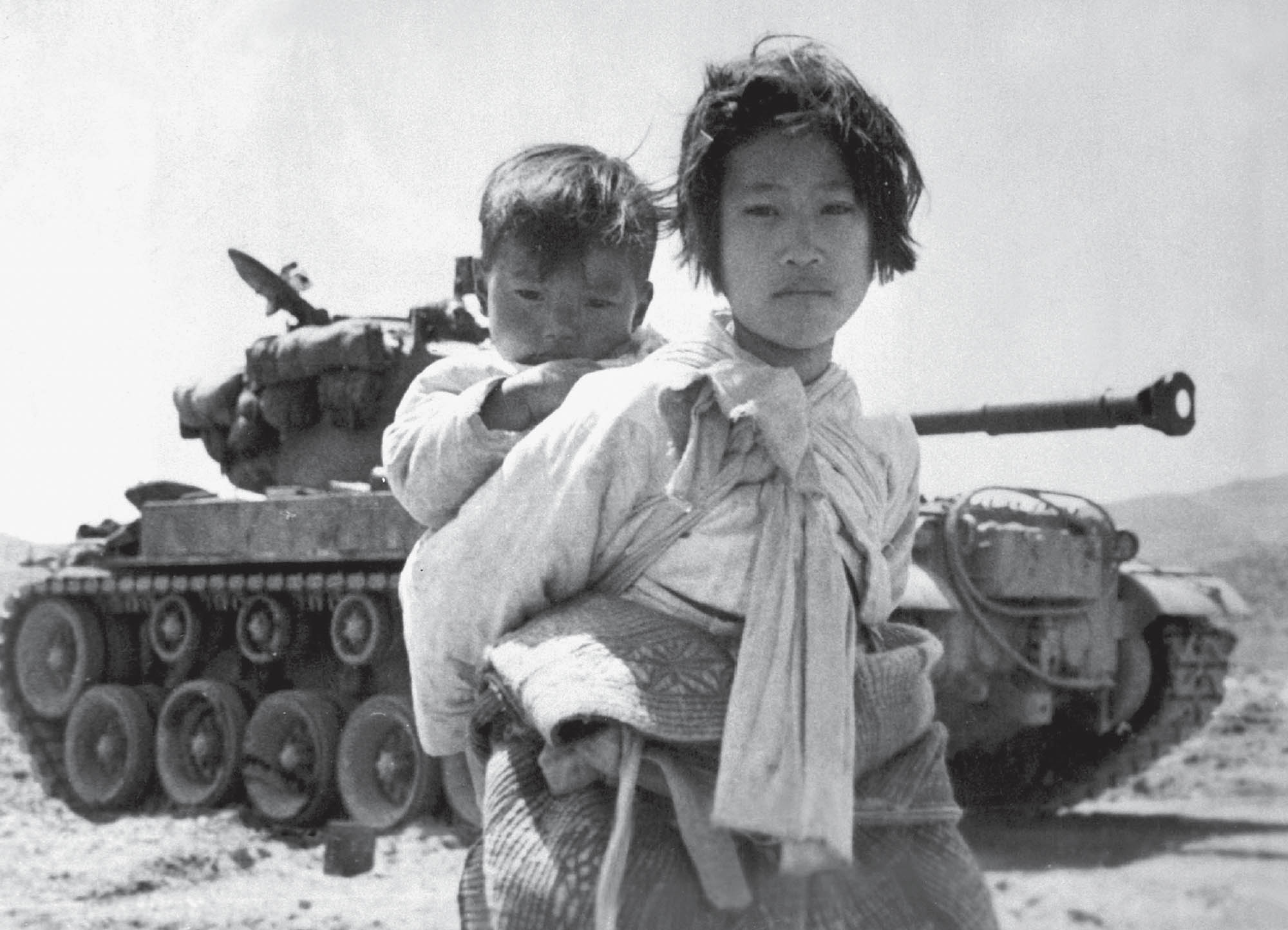 Photo of a young Korean girl carrying a baby boy on her back. Behind them is a military tank.