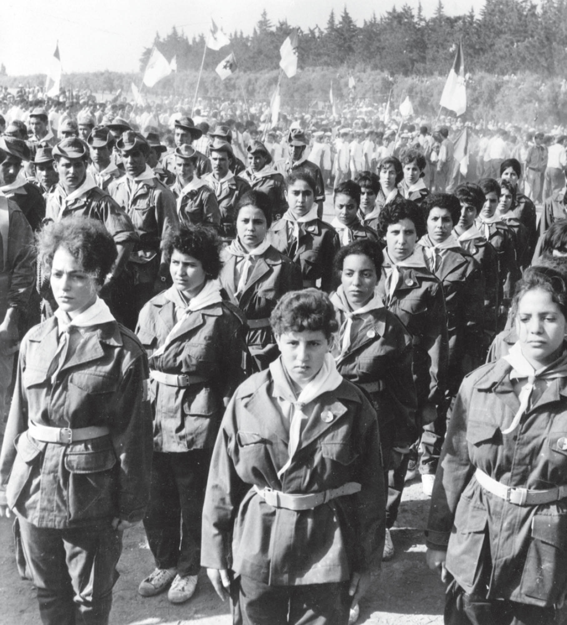 Photo of the women’s section of the Algerian National Army of Liberation standing at attention with their arms fixed at their sides. Each woman is dressed in a military uniform that consists of cargo pants, jackets, belts, and a neck scarf.