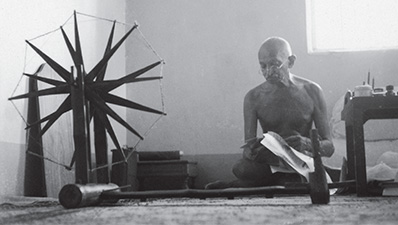 A photograph of Gandhi sitting, cross-legged, in a small room, reading a text. There is a small table on one side of him, and a spinning wheel on the other.