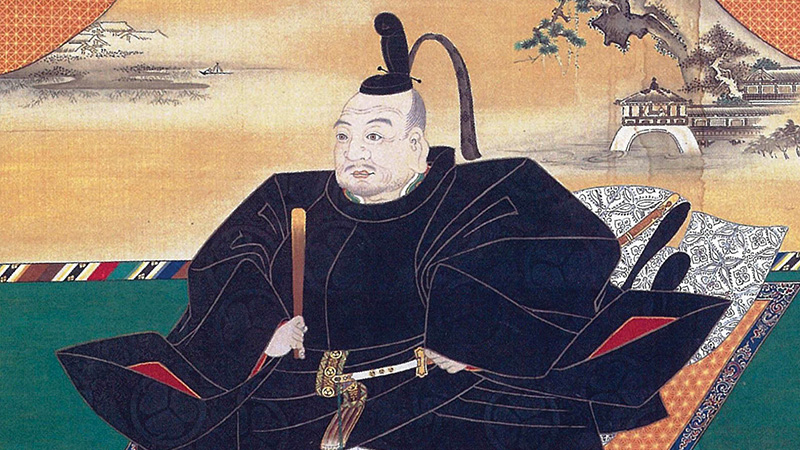 Painting of a Japanese shogun dressed in black robes and sitting cross-legged on an ornate carpet while holding a traditional Japanese paper fan.