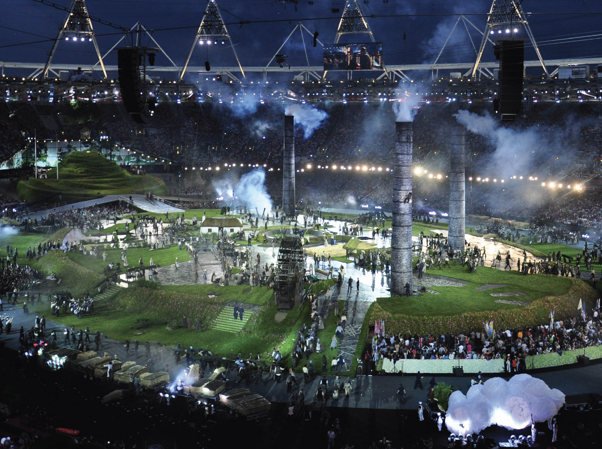 Photo of the inside of a stadium at the opening ceremony of the 2012 Olympics. The stadium has been transformed into an industrial town, complete with three large factory pipes, a water wheel, and groups of people wearing traditional clothing from the era.
