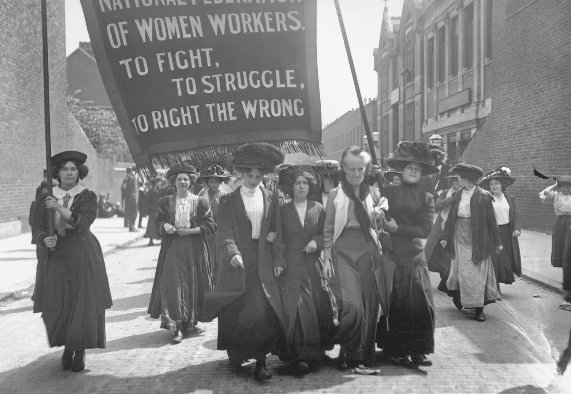 Photo of women’s suffrage protestors marching down a street holding a sign with the slogan, “To fight, to struggle, to right the wrong”.