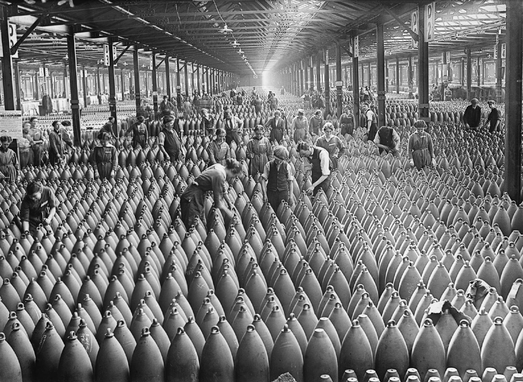 Photo of men, women, and children working in a munitions factory. Rows upon rows of deadly weaponry line the factory floor.