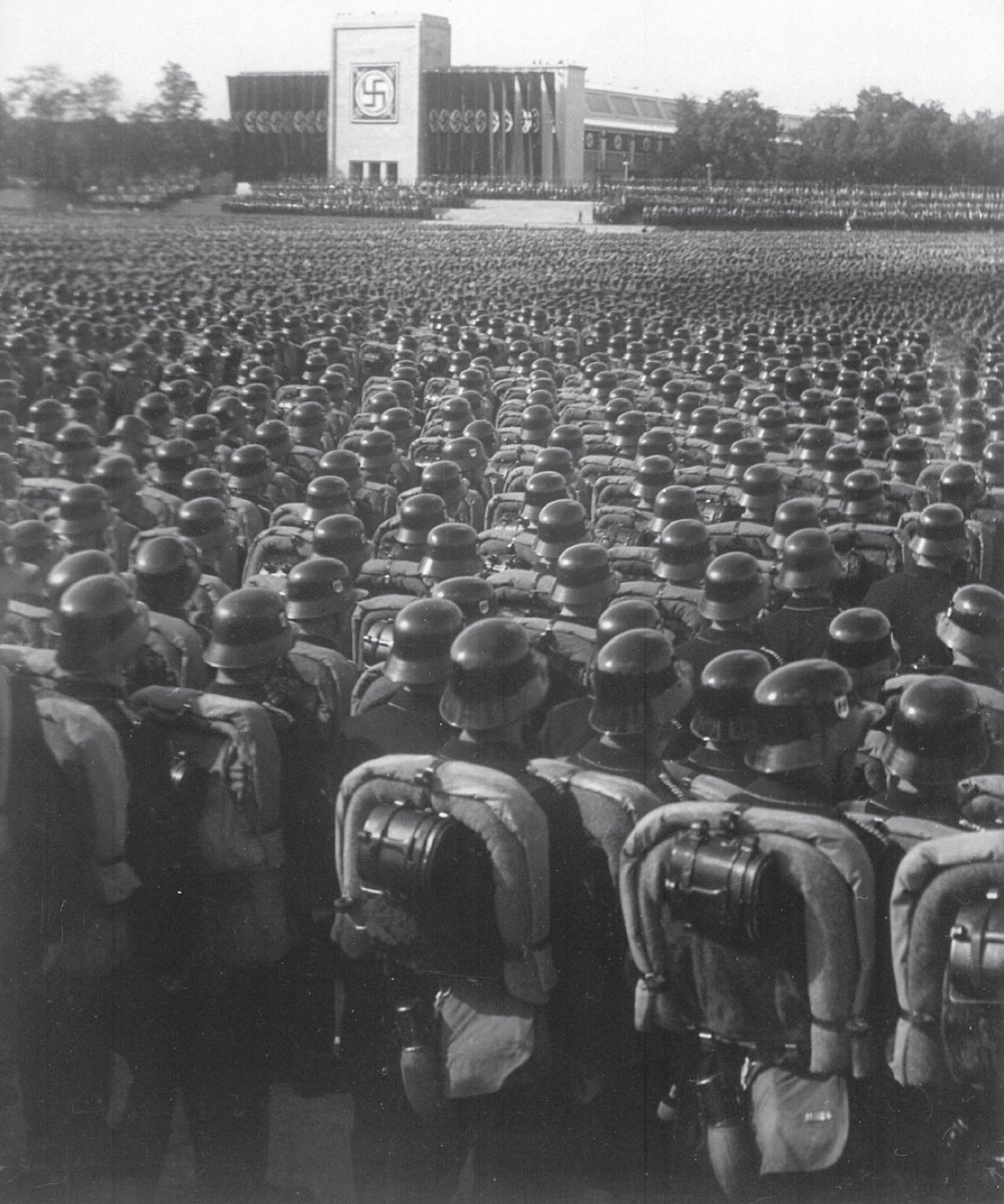 Photo of a crowd of Nazi soldiers who have gathered to listen to Adolf Hitler speak. A large swastika can be seen on the building in the background.   