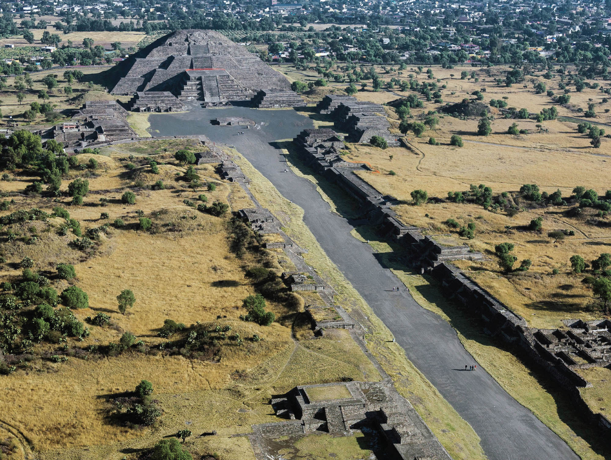 Photo of Teotihuacan, showing a long narrow road to a center area surrounded by the structures that make up Teotihuacan. There is a larger, pyramid structure, and several smaller structures that are leveled and have staircases going up them