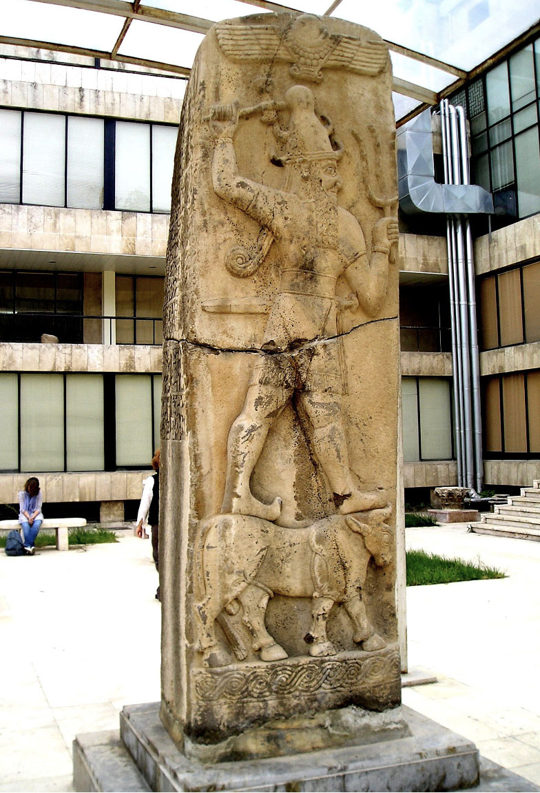 Photo of a pillar carved with the image of a man holding a trident-like weapon and standing on top of a bull