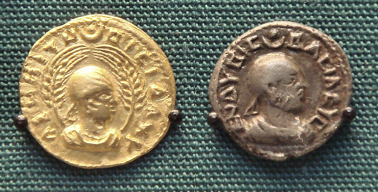 Two gold coins featuring symbols and a depiction of a person’s profile. One is bright and shiny and the other is tarnished, and they feature slightly different markings.