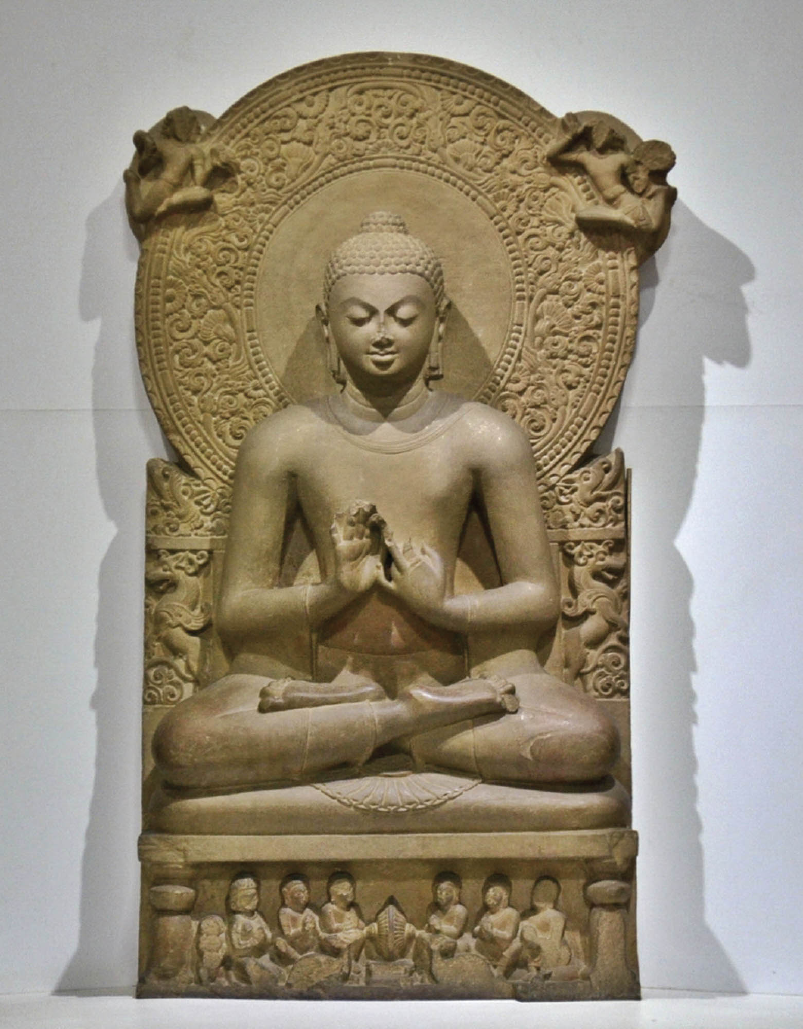 A statue shows the Buddha seated in a cross-legged position with their eyes closed. The Buddha is seated on a throne or chair that is covered in ornate symbols.