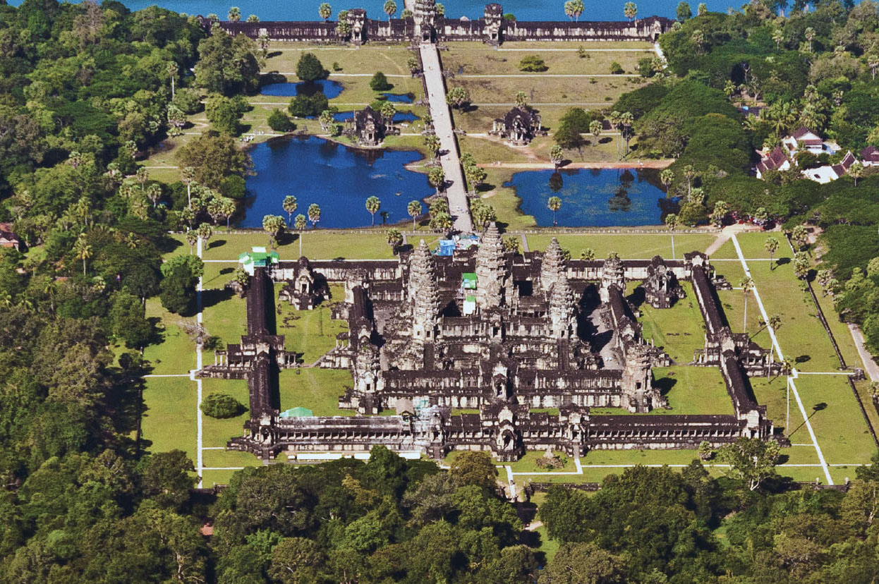Birds-eye view of Angkor Wat, a very large and ornate temple. There is a main structure, surrounded by a wall that borders the temple. Around the temple are blue pools, grass, and trees. 