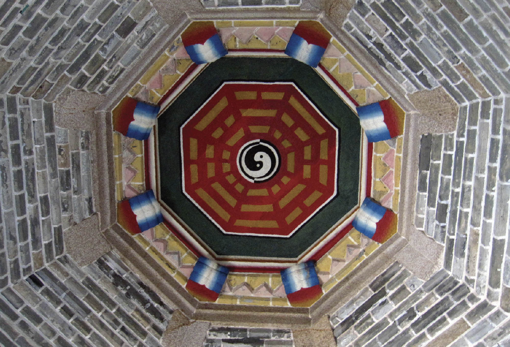 An octagonal ceiling features several symbols in bright colors, surrounding a yin and yang, which is in the center. Leading up to the ceiling are brick walls.