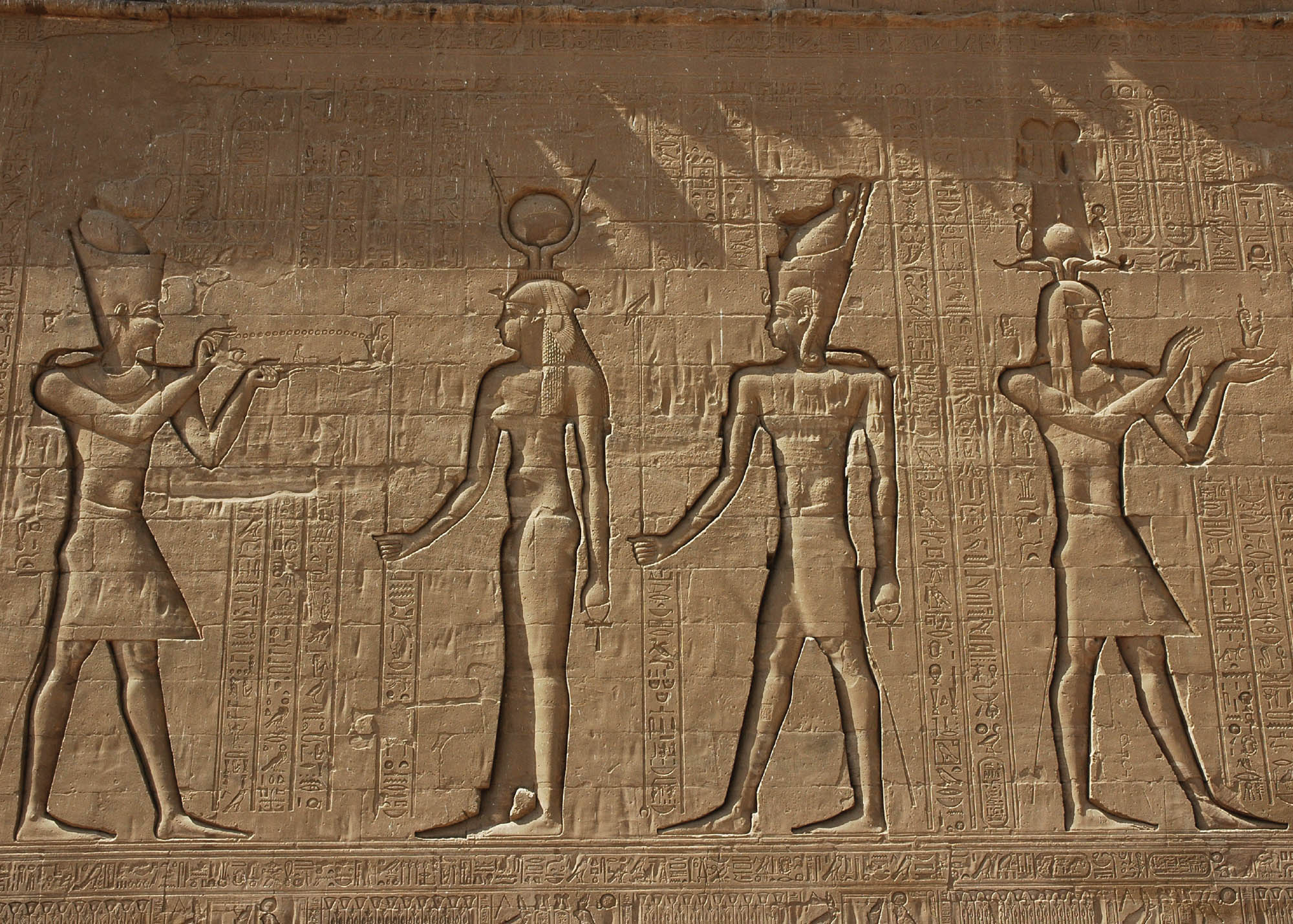 A large stone wall. There is text and symbols that are inscribed in the wall, as well as four figures. One of the figures is holding incense, burning in his hand.