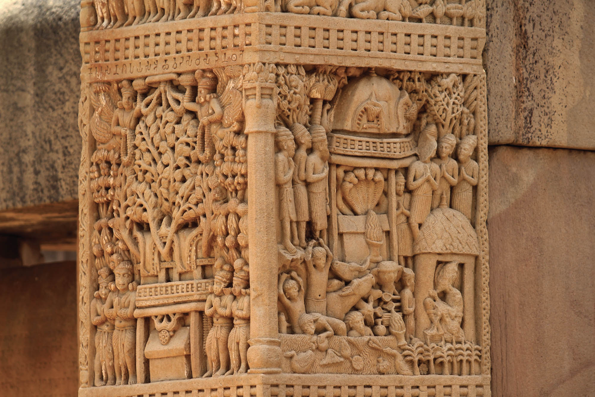 Photograph of a carved stone rectangular pillar. Both sides are carved in great detail featuring many people with their hands together in a prayer position.