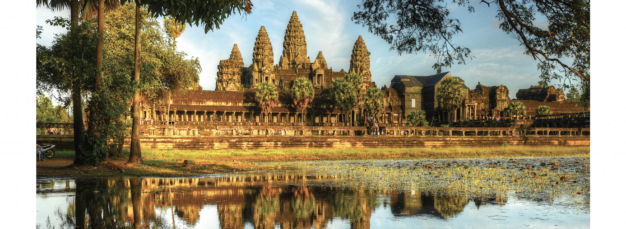 A temple with several ornate domes sits at the edge of a body of water, the reflection of the temple and the surrounding tall trees can be seen in the water.