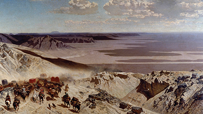 A vast and mountainous desert landscape under blue skies with large white clouds that cast dramatic shadows on the desert below. In the foreground there is a group of people on horses and other animals who have large loads of goods strapped onto them.