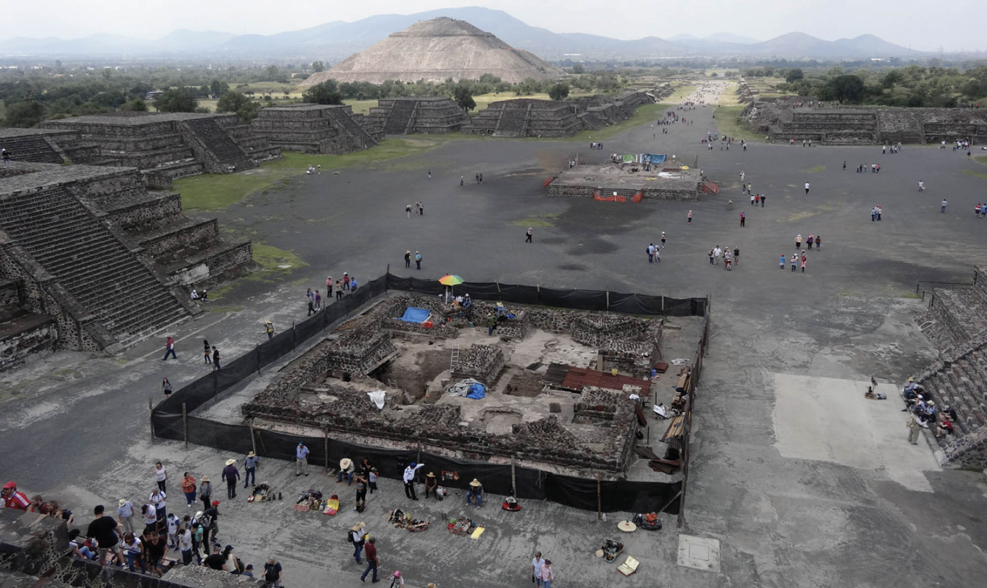 In the center of the city of Teotihuacan, people crowd around an archaeological site. Inside the site we can see remnants of brick structures. The site has a black fence around it.