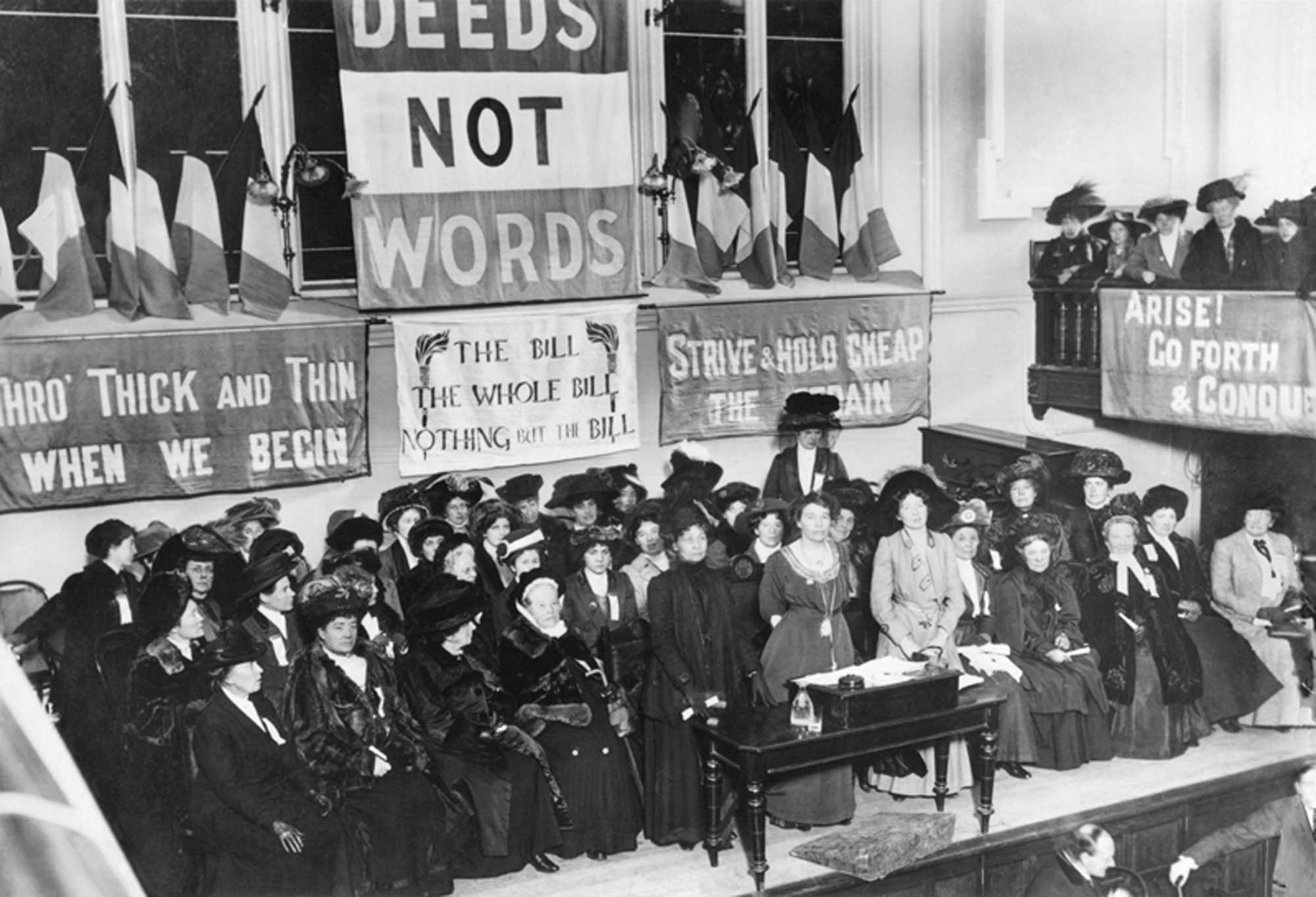 A photograph of a suffragette meeting. Women stand in a large meeting room and hold up banners. One reads, “Arise! Go forth & Conquer”.