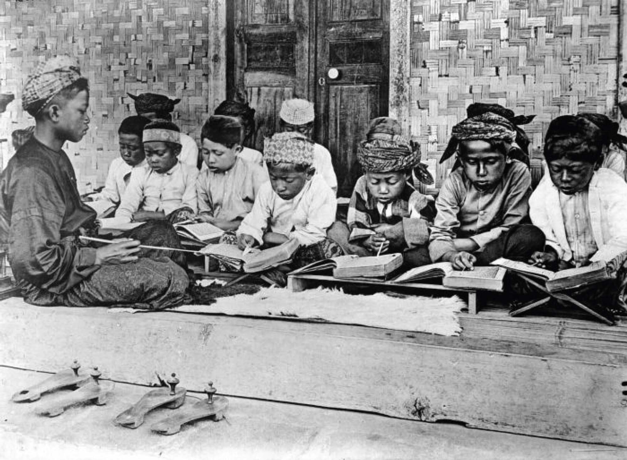 A photograph of a group of young children, seated cross-legged. They are sitting outside, studying.